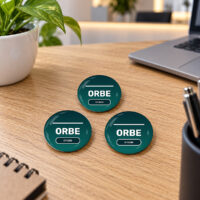 Três bottons personalizados de 5,8 cm com identidade visual corporativa sobre uma mesa de trabalho