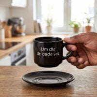 Mão segurando a xícara preta personalizada com a frase Um dia de cada vez, com o pires na mesa, mostrando o tamanho real